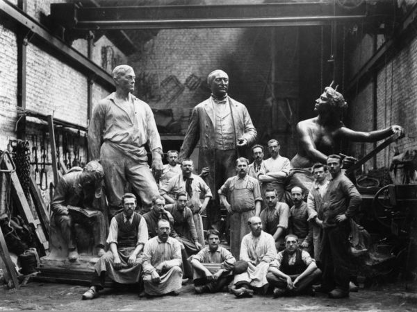 Ancienne photo en noir et blanc datant de 1900. Des ouvriers de la Compagnie des Bronzes posent dans la halle de montage devant trois grandes statues en bronze.