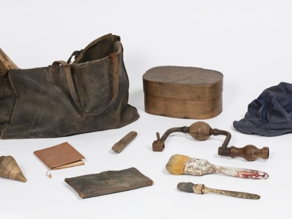 Musette en cuir d'un ouvrier plombier contenant une casquette,deux pinceaux, un burin en bois,un vilebrequin en bois et acier, une toupie ,un carnet, une pochette, un manche en bois et une boîte à tartines en bois.