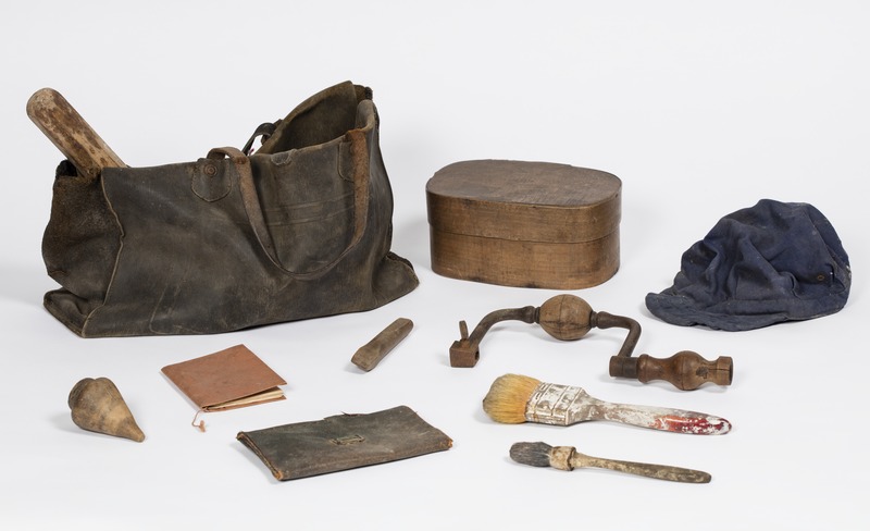 Musette en cuir d'un ouvrier plombier contenant une casquette,deux pinceaux, un burin en bois,un vilebrequin en bois et acier, une toupie ,un carnet, une pochette, un manche en bois et une boîte à tartines en bois.